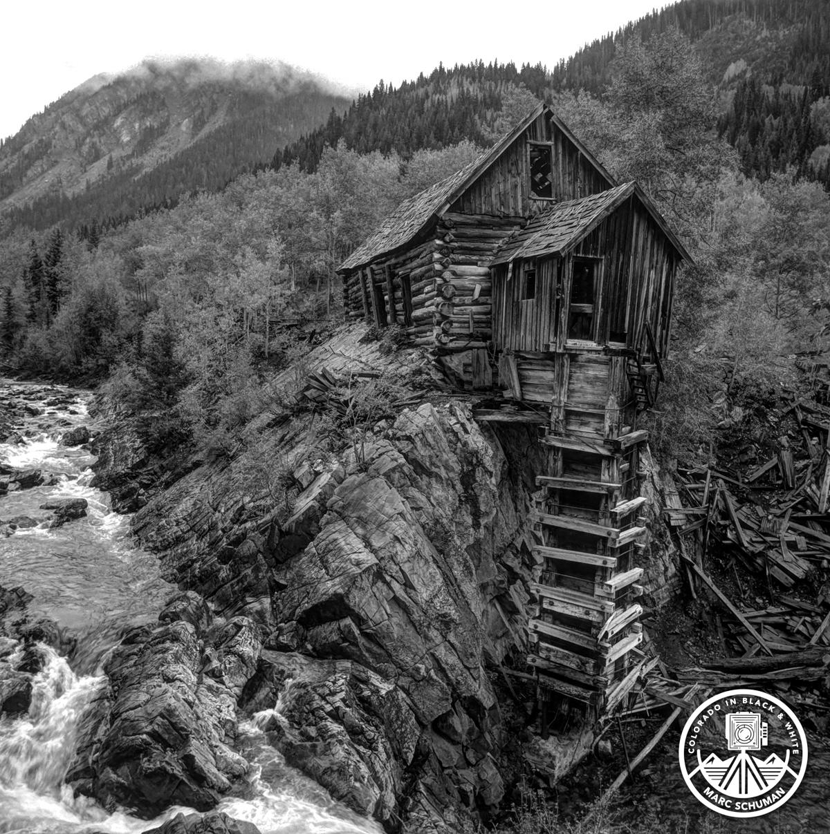 Black and white photograph of the Crystal Mill, a weathered wooden mill perched on a rocky outcrop above the rushing Crystal River in Colorado, with aspen-covered mountains behind