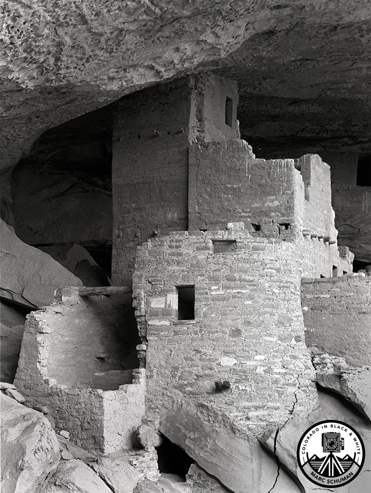 Black and white vertical photograph of the stone Round Tower in the Cliff Dwellings at Mesa Verde, Colorado, sheltered under a rock overhang