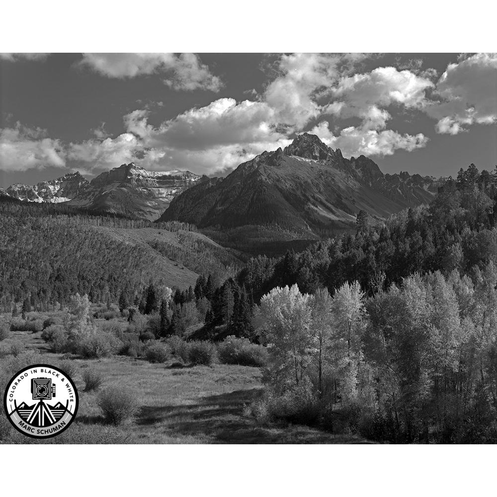 Black and white photograph of Mt. Sneffels in the San Juan Mountains of Colorado, with large clouds over the peak and a meadow and stands of aspen and cottonwood in the foreground
