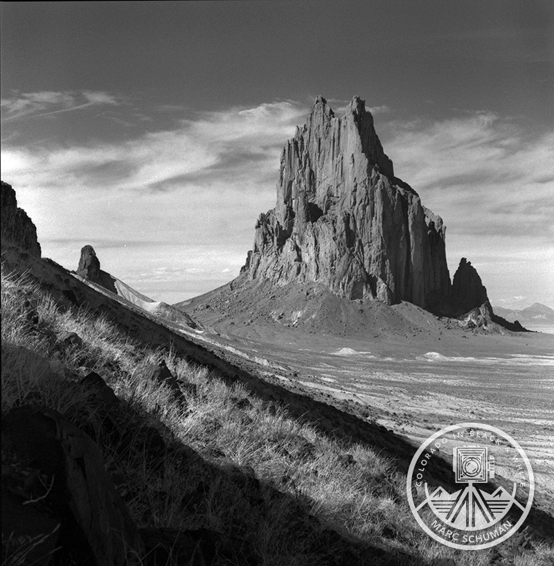 Black and white photograph of Shiprock rising from the New Mexico desert, seen from a steep grassy slope under streaks of cloud